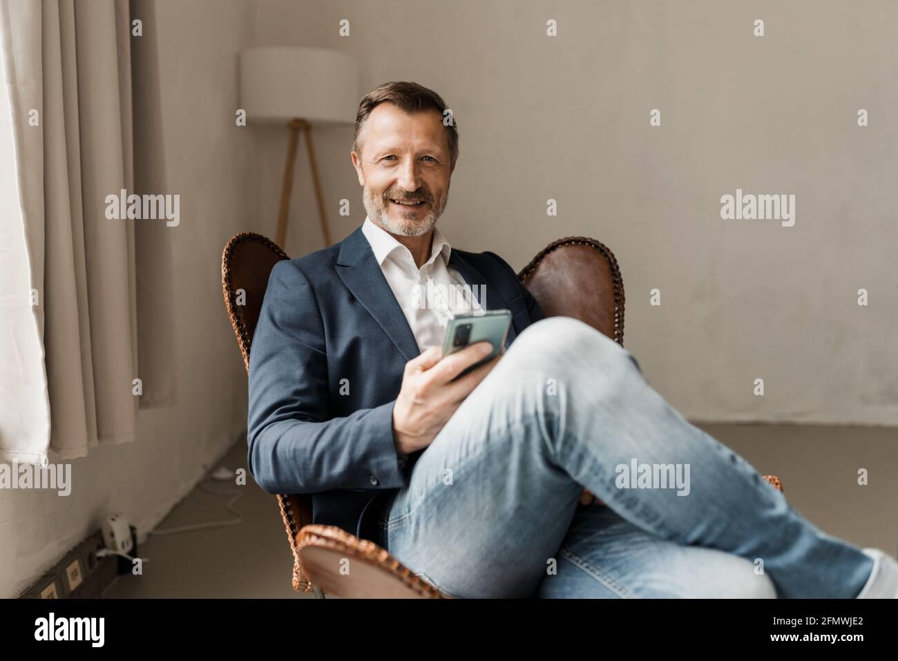 Attractive modern middle-aged man in jeans and stylish jacket relaxing in a leather chair with mobile phone in hand smiling at the camera Stock Photo