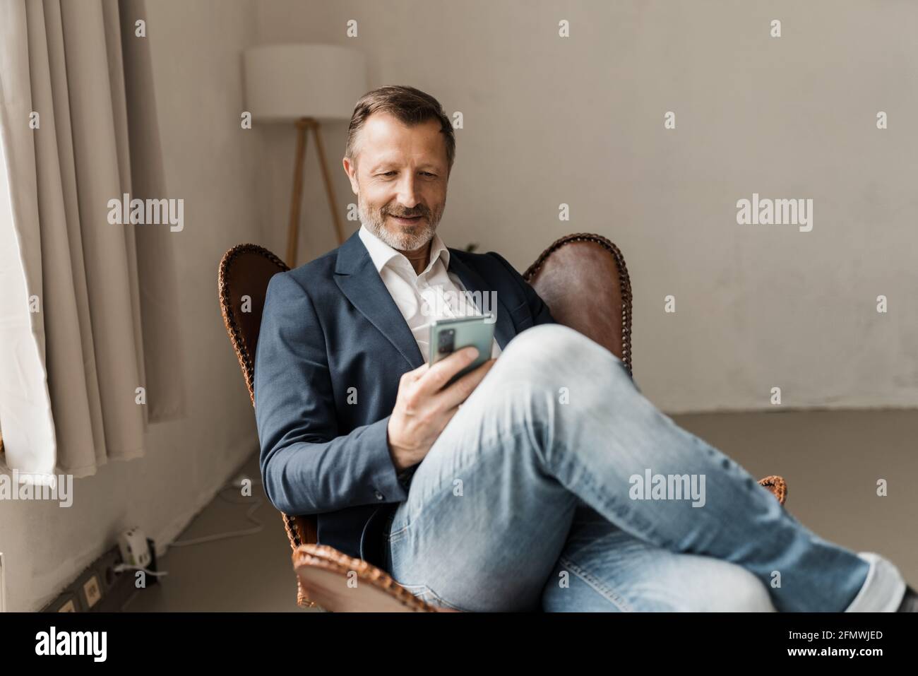 Modern trendy businessman relaxing in a leather chair reading a message or watching media on his mobile phone with a quiet smile Stock Photo