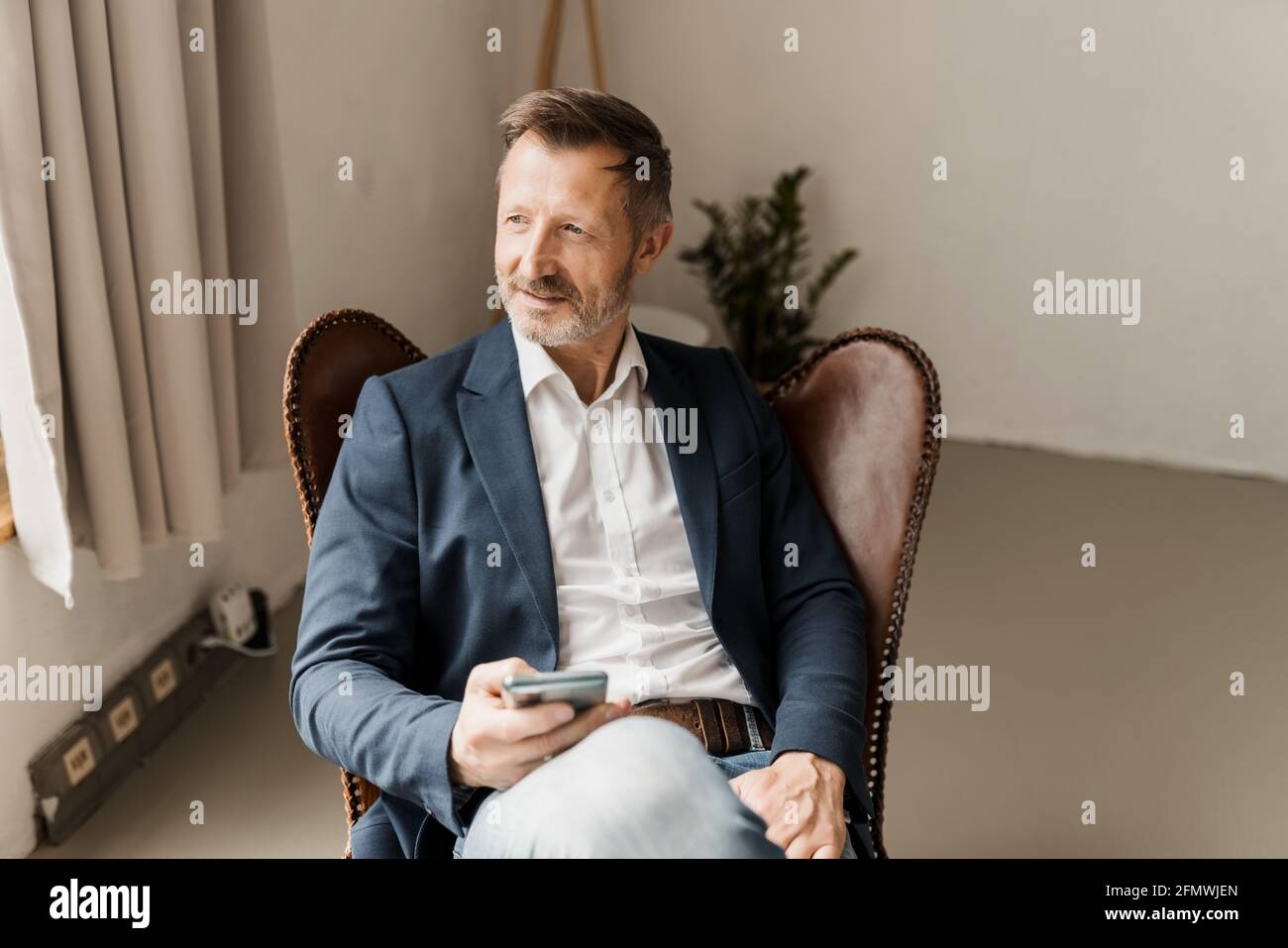 Smart relaxed businessman sitting staring through a window with an intrigued interested expression as he sits in a comfy chair with his mobile phone Stock Photo