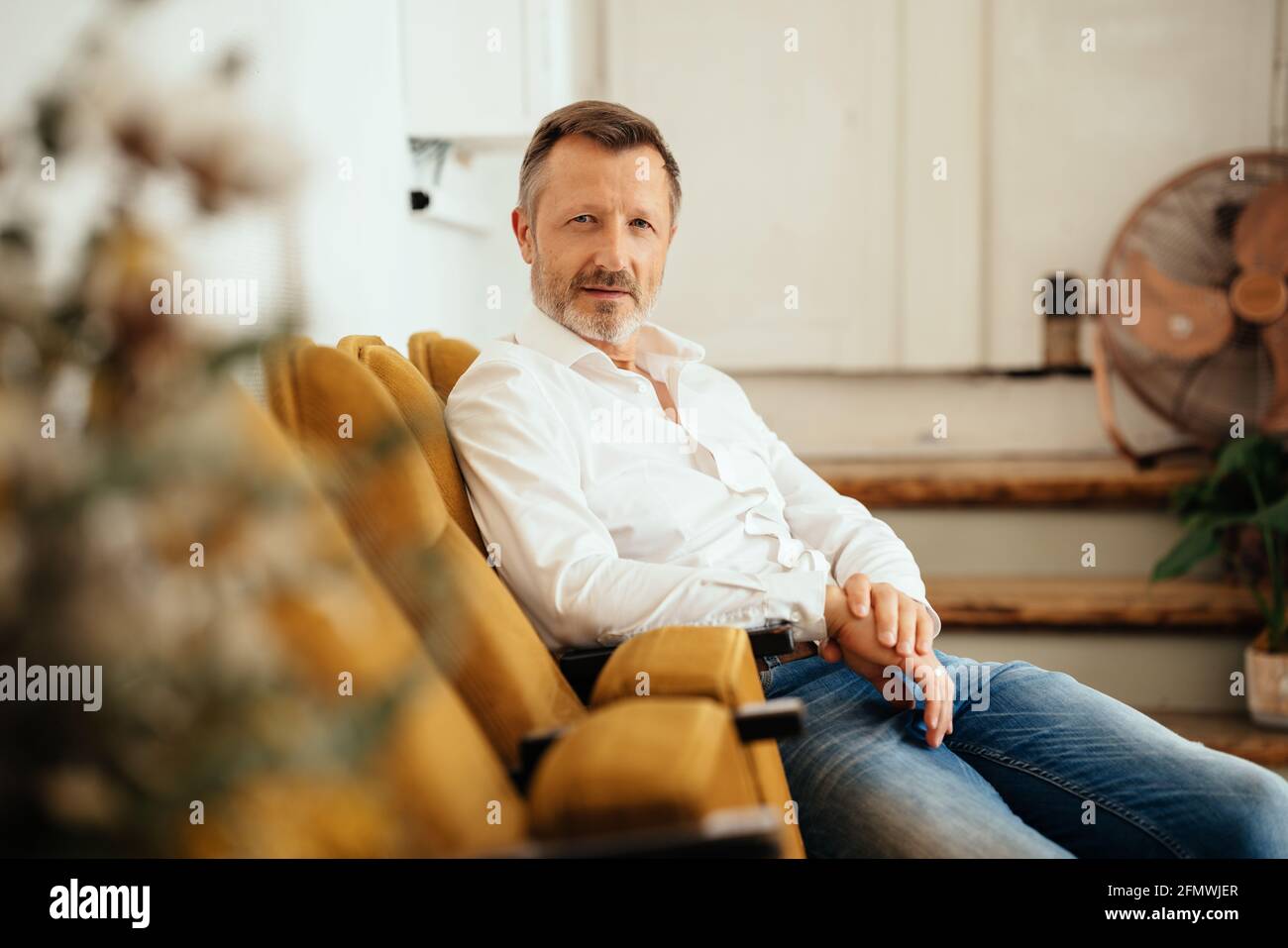 Middle-aged man relaxing in chair staring intently at the camera with an inscrutable expression indoors viewed past a potted plant with copyspace Stock Photo