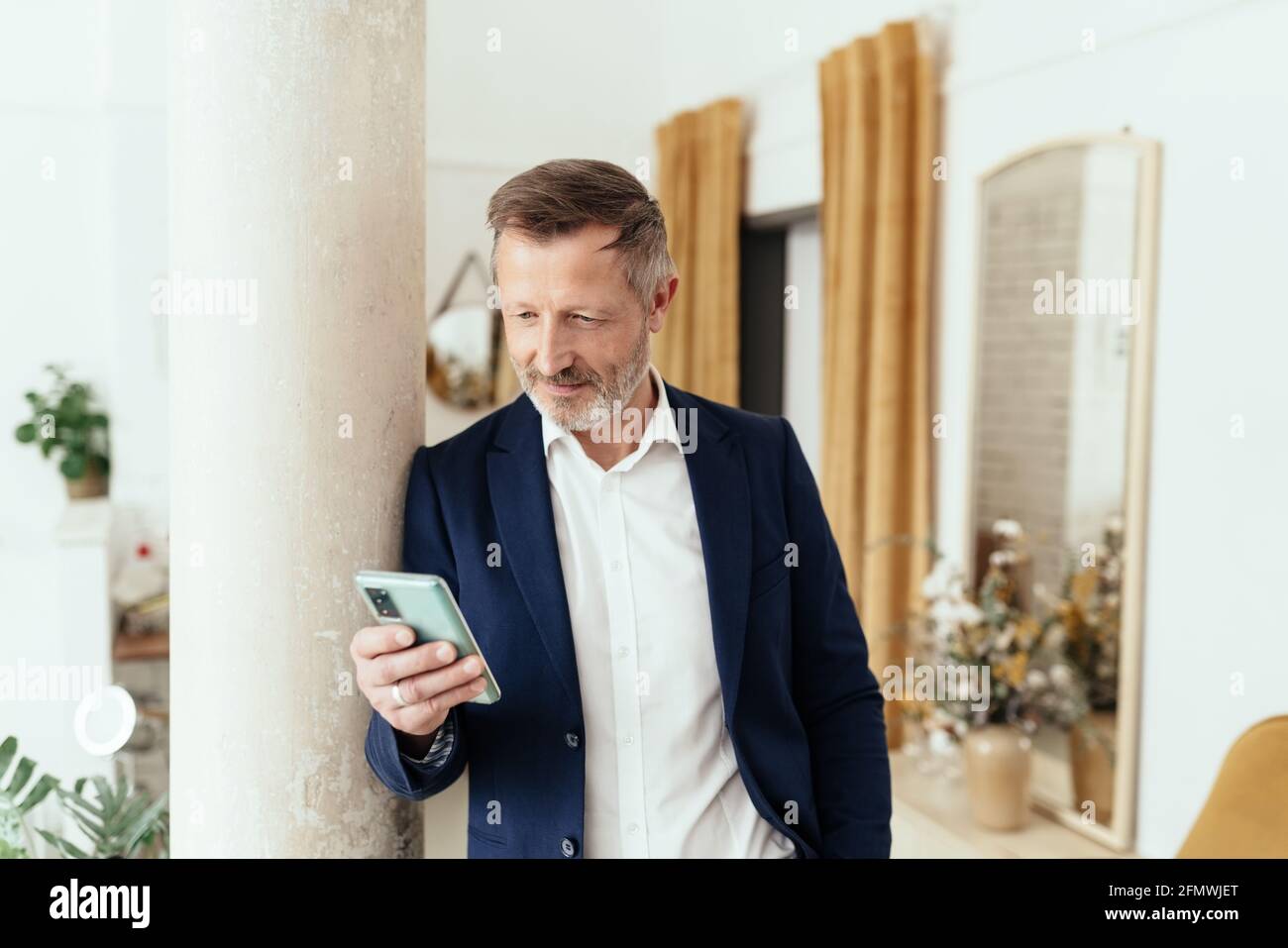 Relaxed businessman in jacket standing leaning against a pillar reading a phone message on his smartphone with a thoughtful look of concentration indo Stock Photo