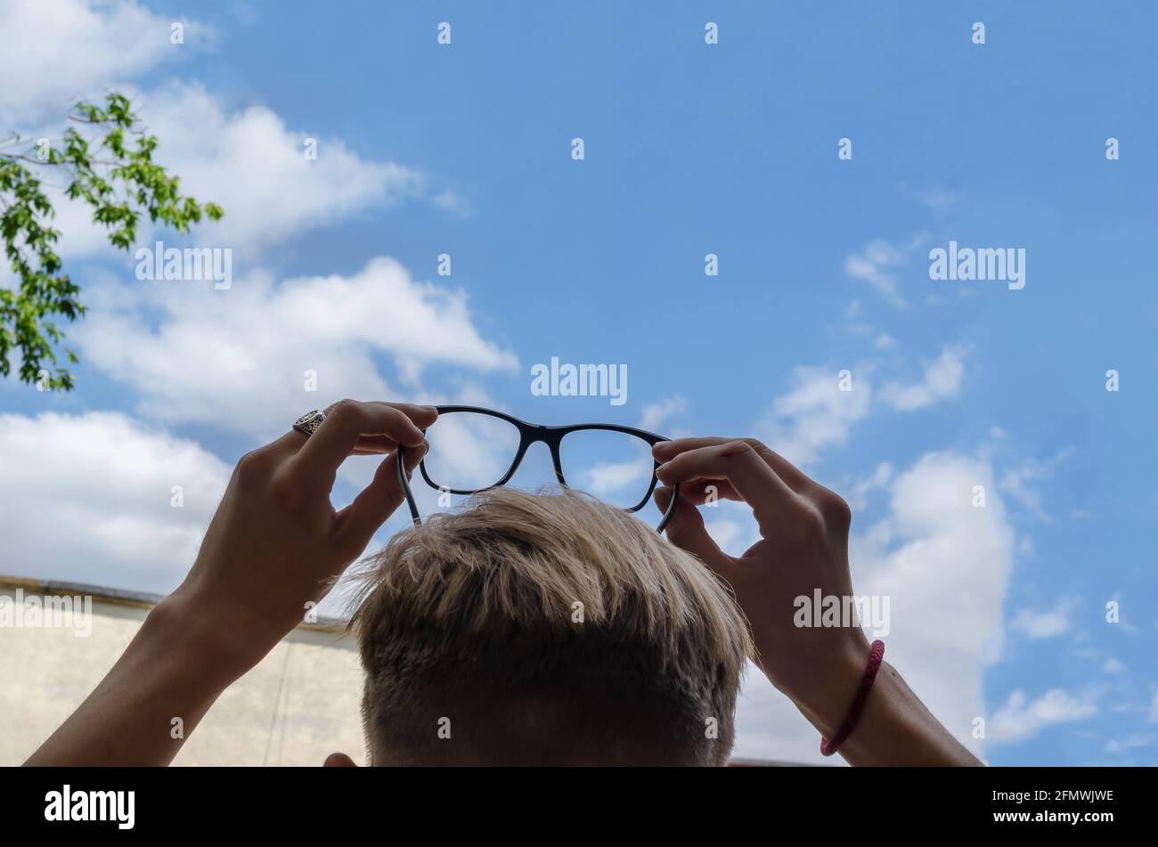 A blond man looks up at the sky holding his glasses over his head. The young