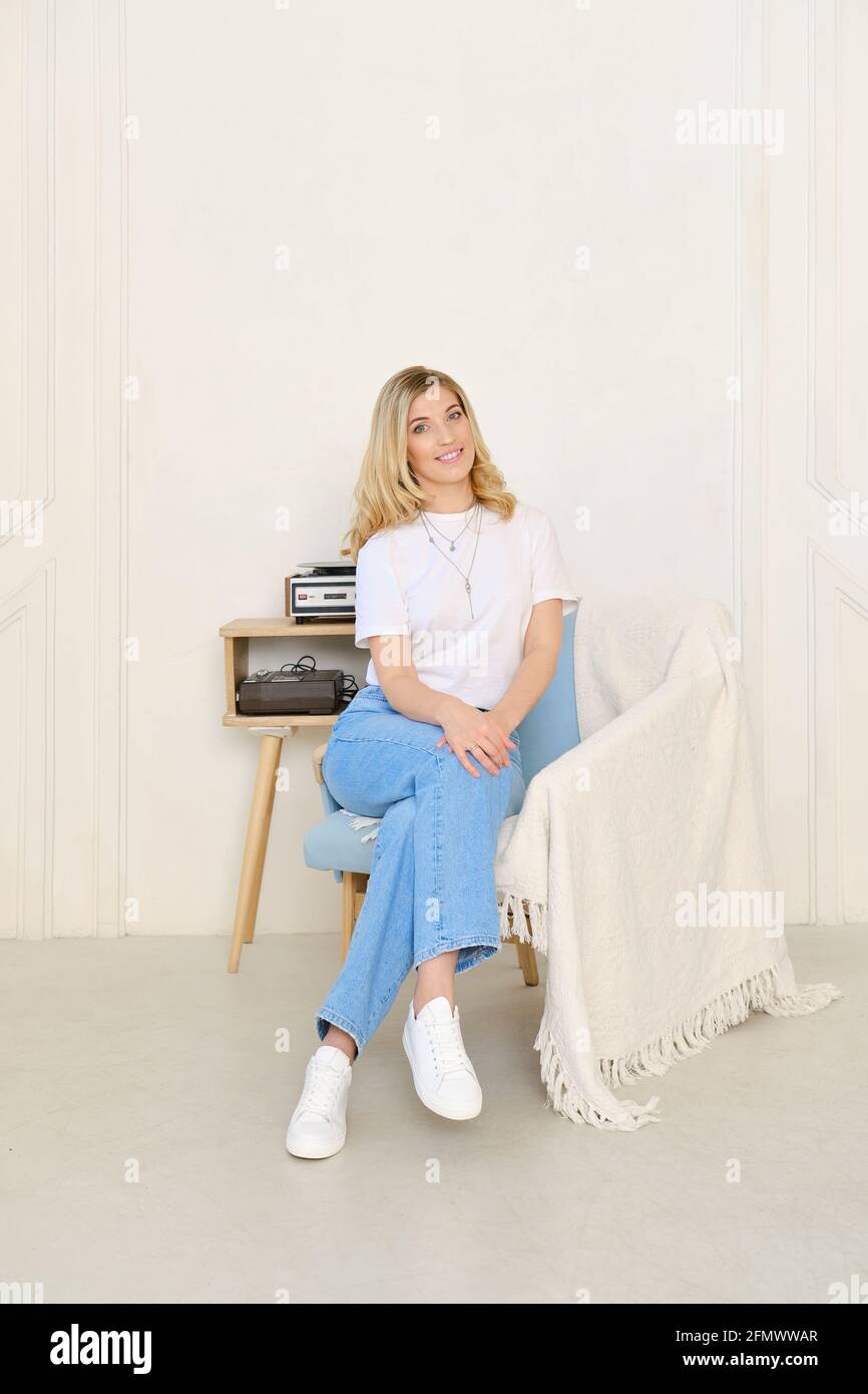 Woman in wide jeans and t-shirt sits in armchair covered with plaid and listening to the music Stock Photo