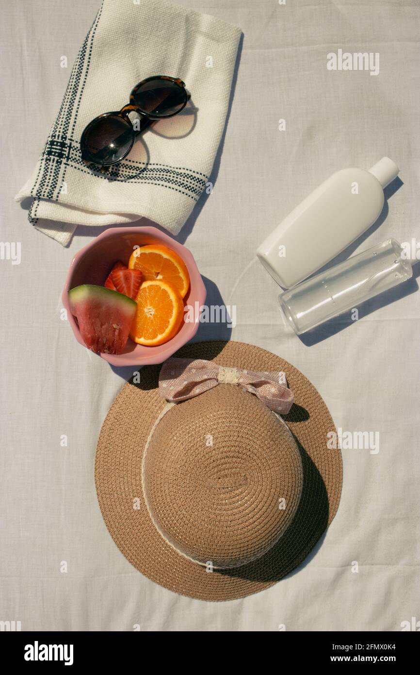 Vertical image of fruits in a pink bowl together with a hat, sunglasses, sun cream and hydrating water on a white fabric background. Summer 2021. Stock Photo