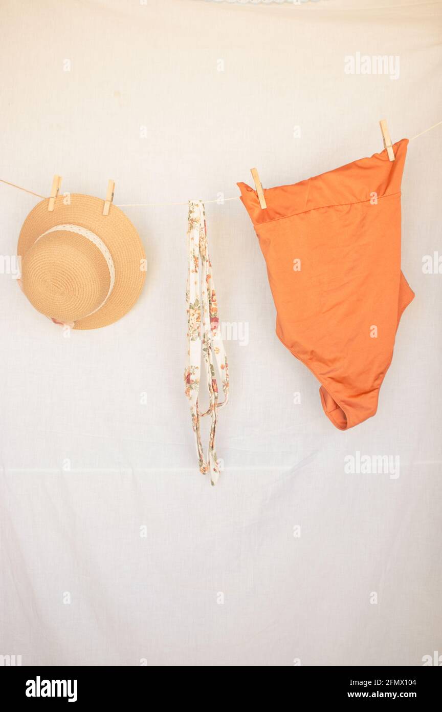 Vertical image of a summer hat, a belt and an orange swimsuit on a clothesline on a white isolated background. Summer Concept 2021. Stock Photo