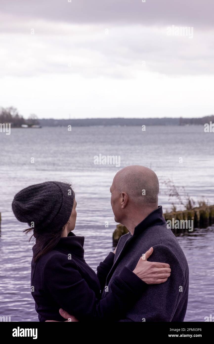 couple in black coats holding each other and looking at lake Stock Photo