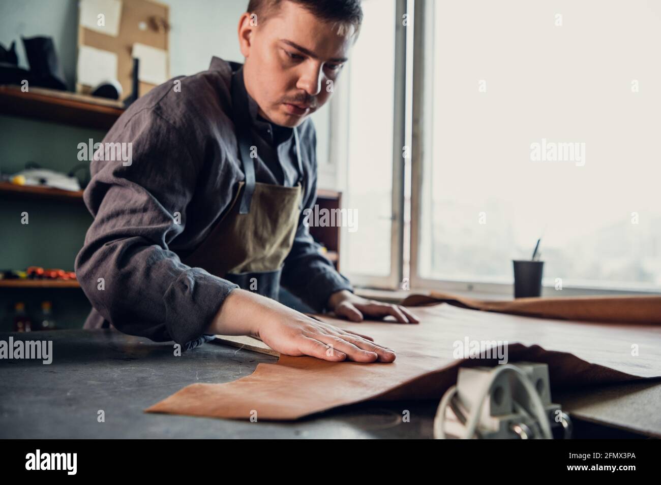 A young apprentice in a boot workshop prepares leather for further use on a large table. Stock Photo