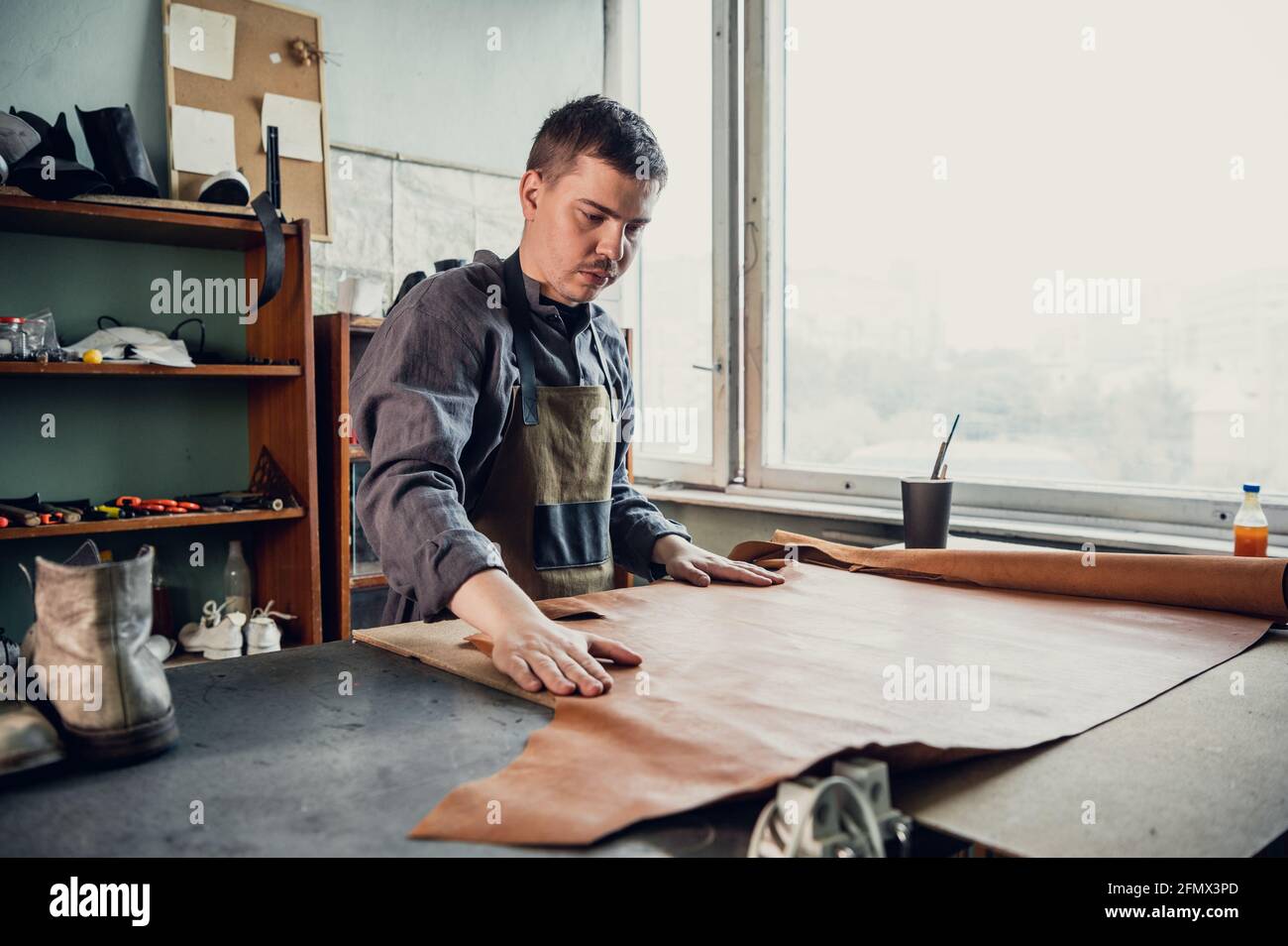 A young apprentice in a boot workshop prepares leather for further use on a large table. Stock Photo