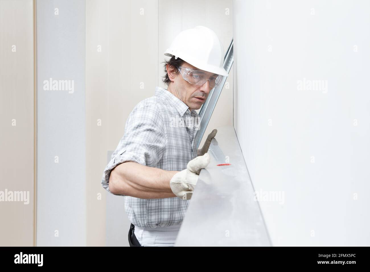 Man drywall worker or plasterer checking level of white plasterboard wall with bubble level at construction site. Wearing white hardhat, work gloves a Stock Photo