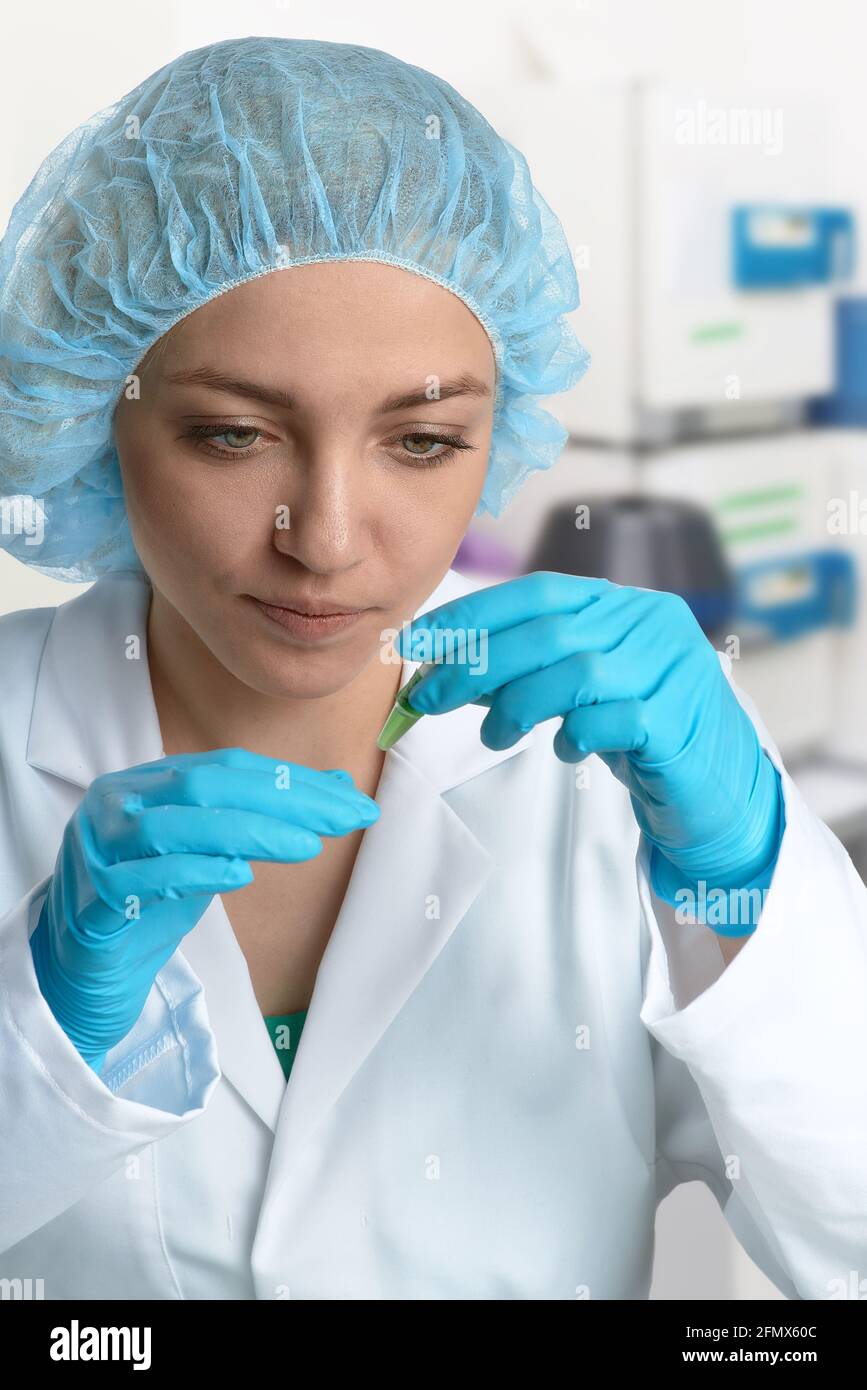 Pharma female tech works in laboratory. Caucasian young woman in protective gloves, hat and white gown pipettes sample in vial. Lab interior Stock Photo