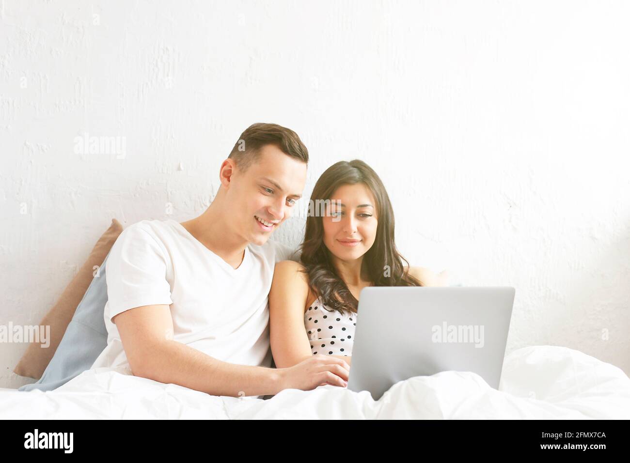 Couple in bed with laptop. Young american man & woman, husband & wife in love holding staring pc notebook computer. Male & female model browsing surfi Stock Photo