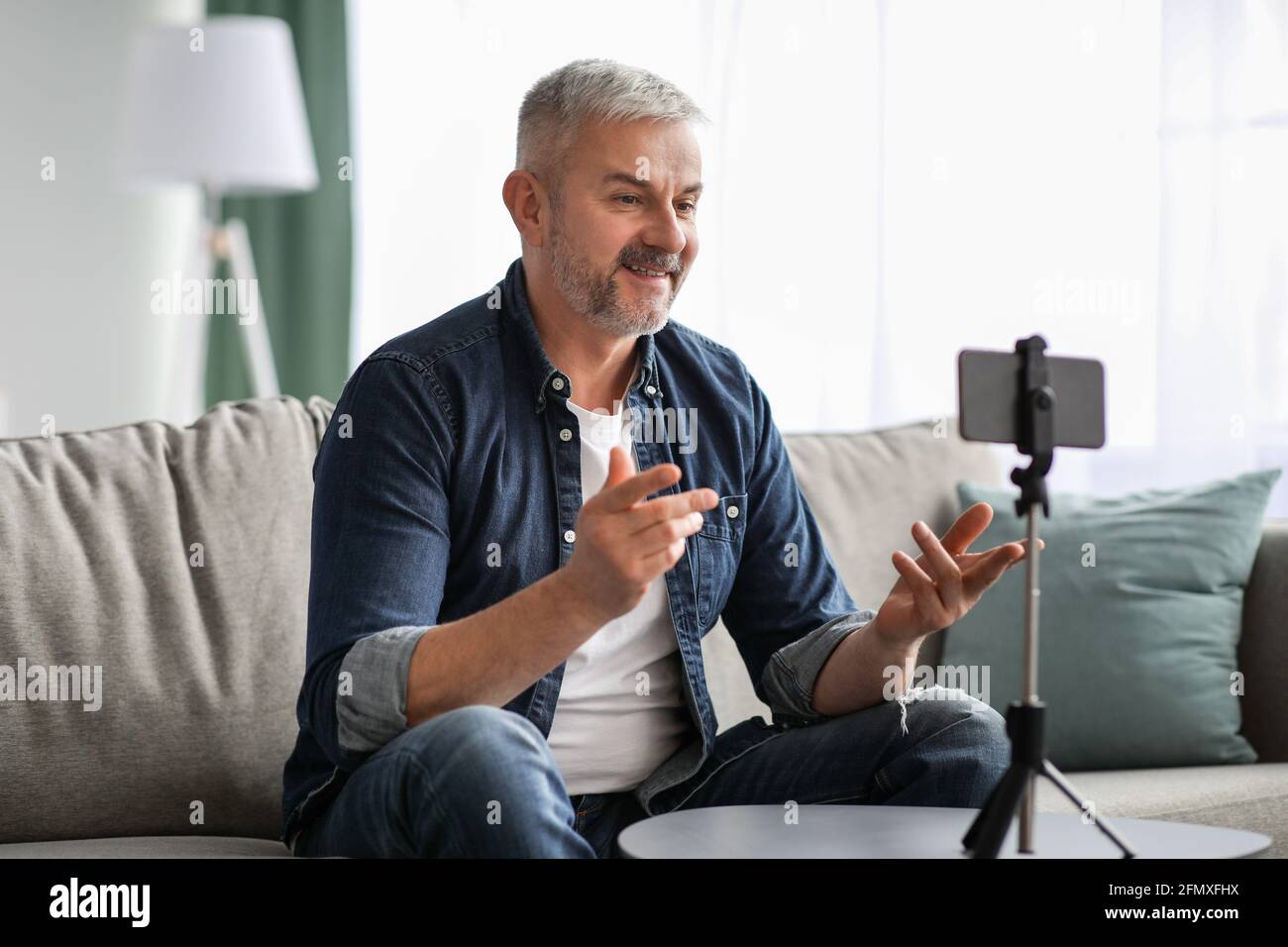 Attractive mature man broadcasting from home, using smartphone on tripod Stock Photo