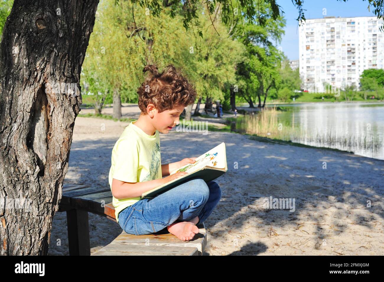 A cute little boy sits on the shore of the lake and reads an old book. Stock Photo