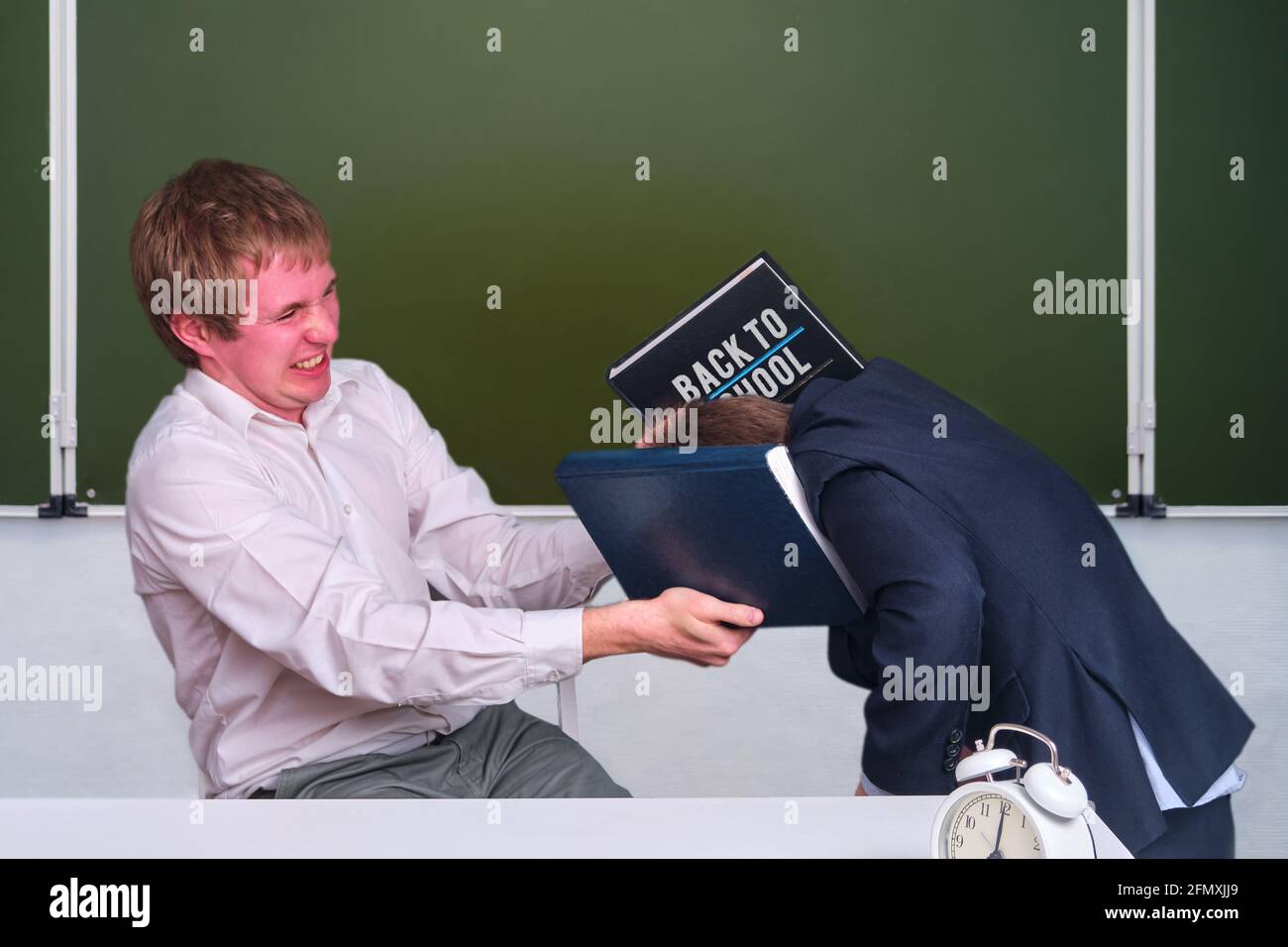 Angry teacher beats a schoolboy with a book in the classroom at the blackboard, copy space Stock Photo