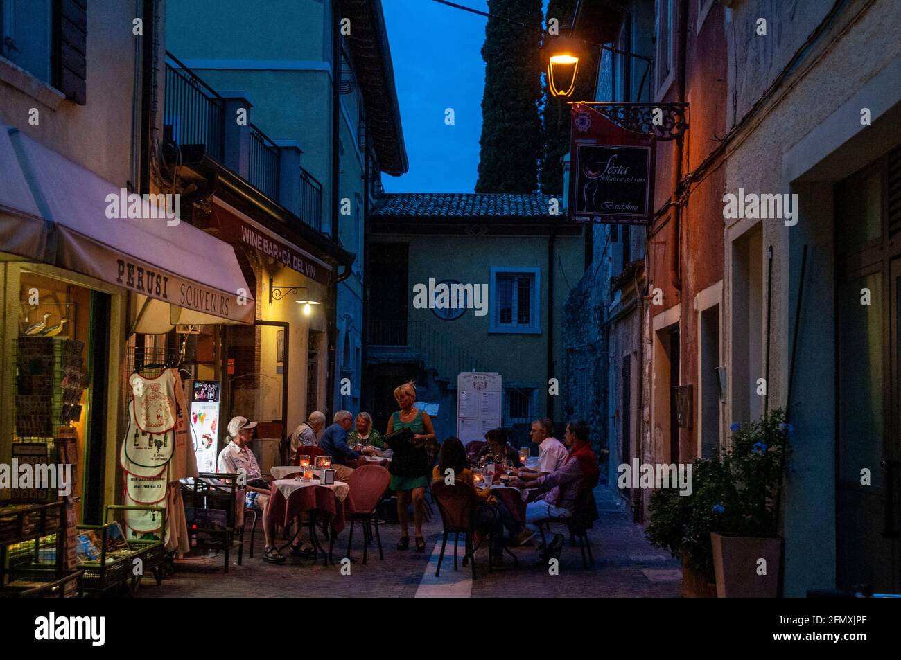 A mix of drinkers and diners sitting at a bistro, lit by street lamps in the early evening at Bardolino, on the eastern shore of Lake Garda in the Ven Stock Photo