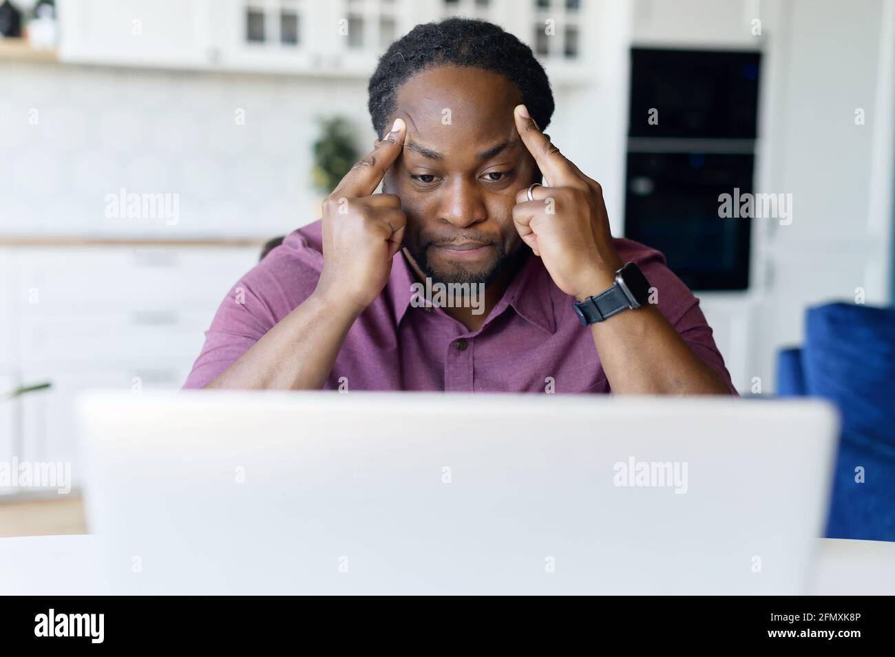 Concentrated African male freelancer using laptop for remote work at home, dark skinned man absorbed with reflections, solving difficult business tasks, staring at the screen focused Stock Photo