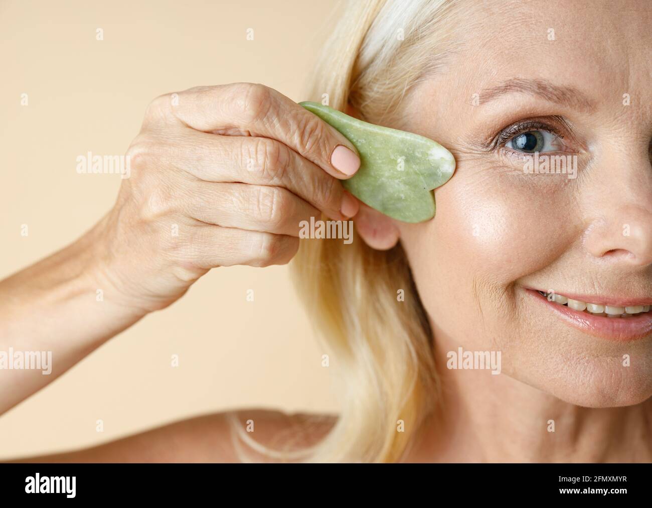Cropped portrait of mature blonde woman with perfect glowing skin looking at camera, holding jade Gua Sha massage tool for skin care and beauty Stock Photo