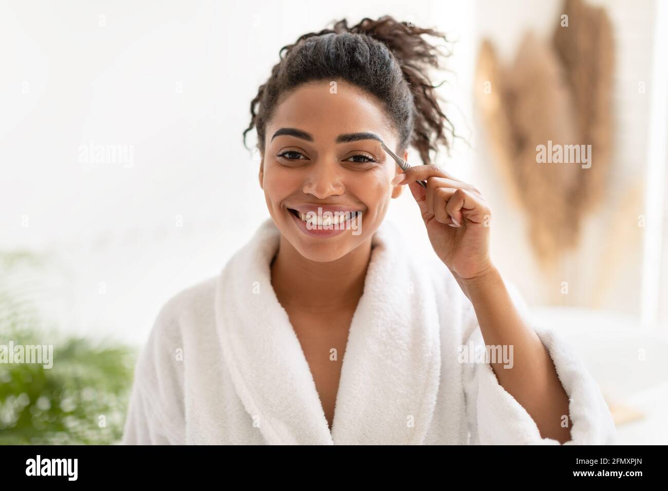 African American Woman Styling Plucking Eyebrows With Tweezers In Bathroom Stock Photo