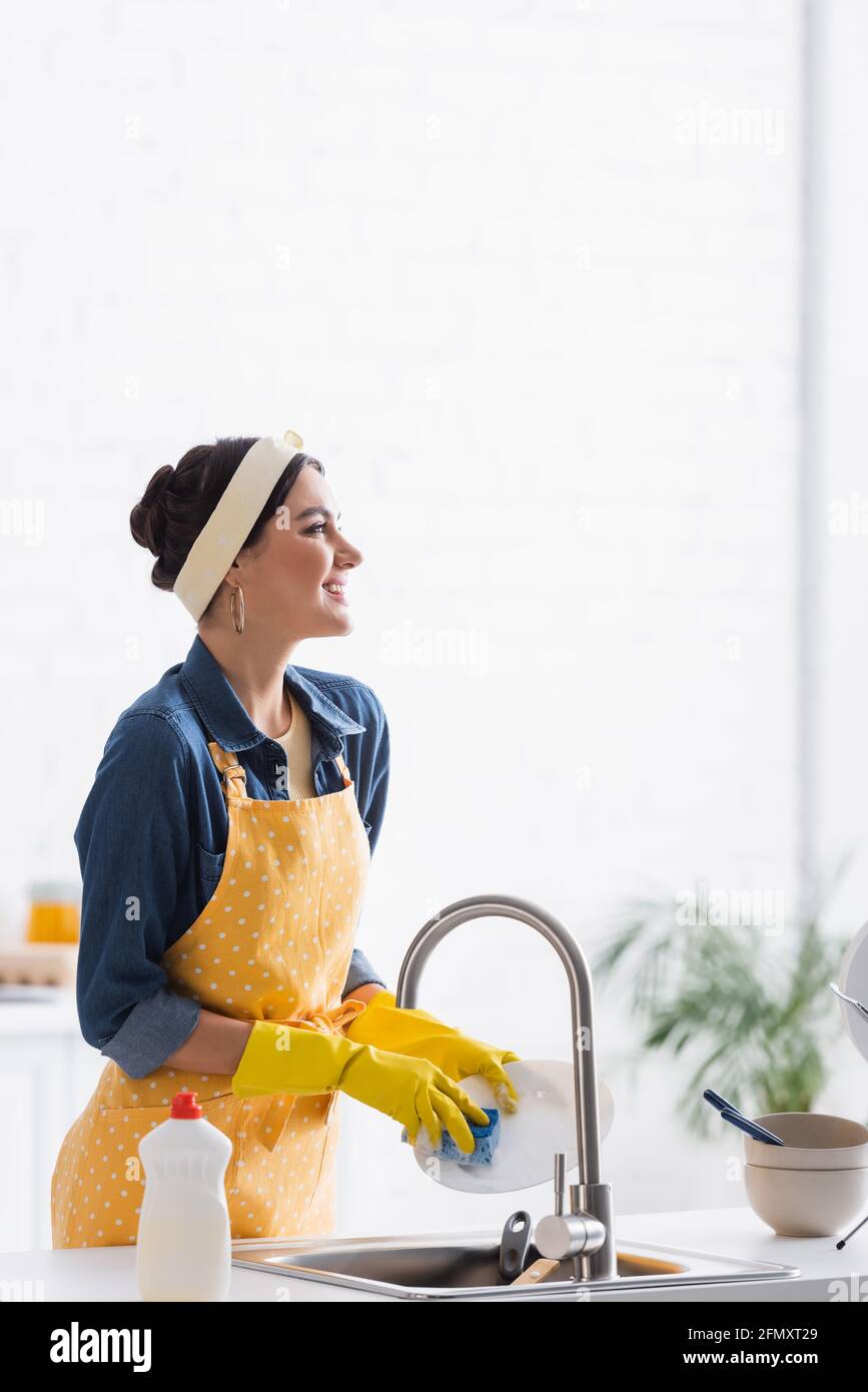 Cheerful woman in apron and rubber gloves washing plate in kitchen ...