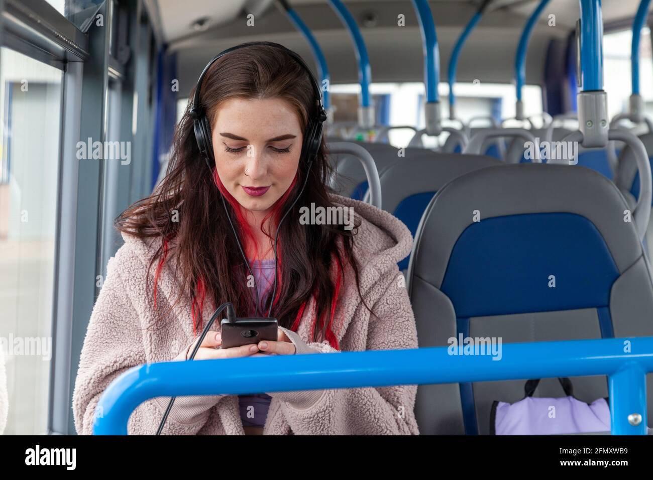 A young woman sitting on a bus with headphones on and looking at her phone Stock Photo