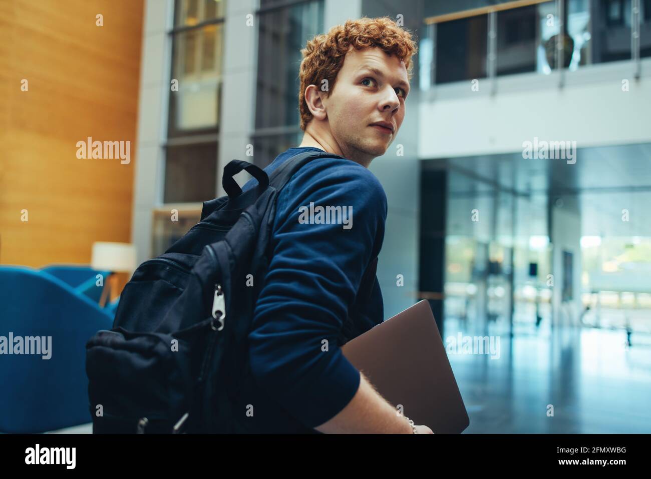 college student carrying his bag and laptop in campus. Young man turning back over his shoulder and walking in college campus. Stock Photo