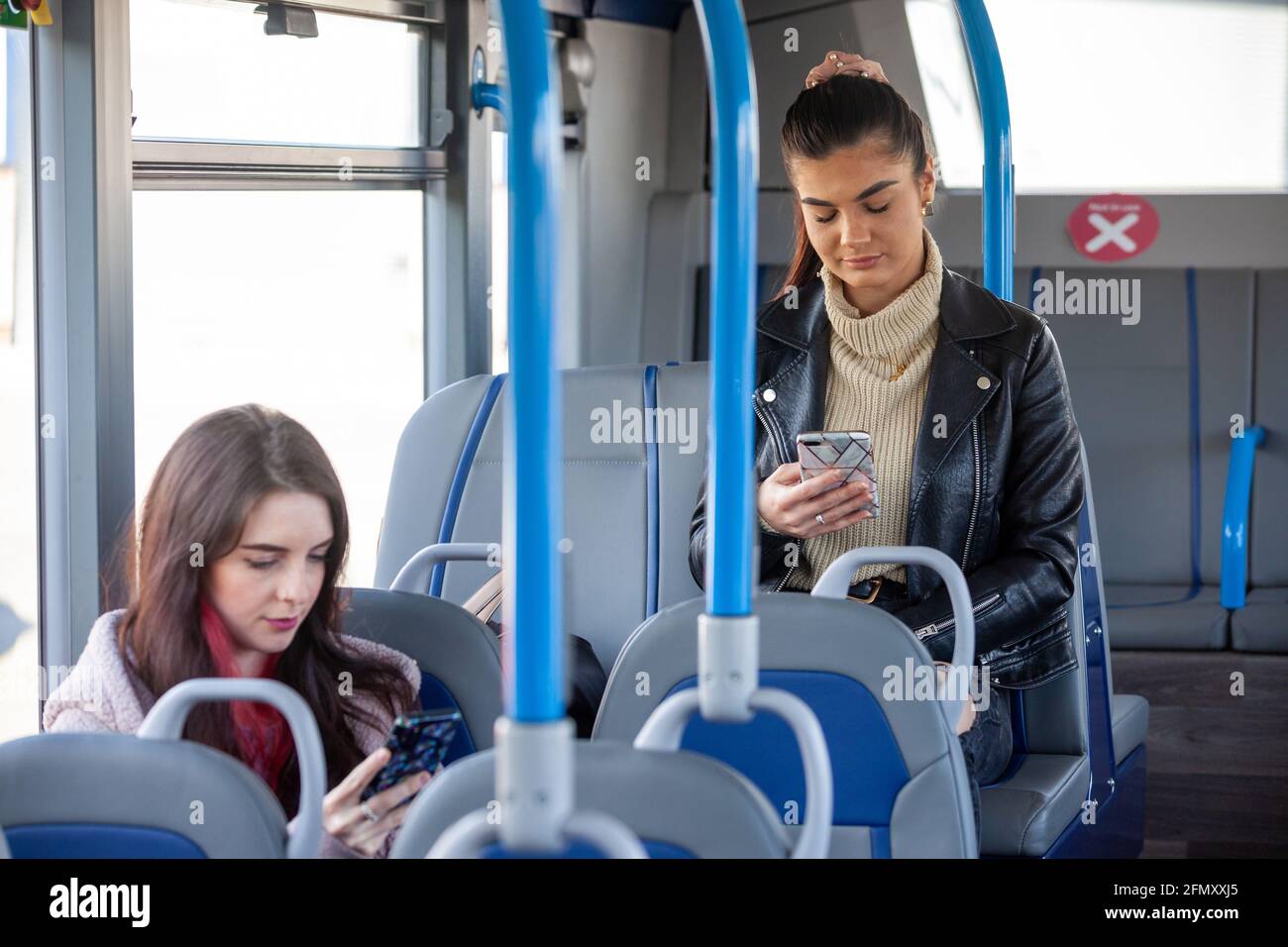 Two women in their 20's travelling on a bus and looking at their phones. Stock Photo