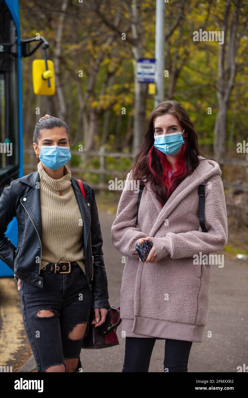 two young women wearing face coverings and waiting in front of a bus at a bus stop. Stock Photo