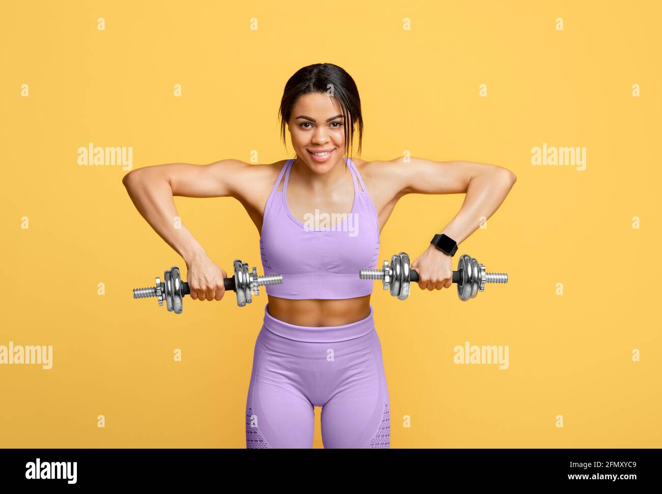 Arms workout. Happy young black lady doing training with two bumbbells,  standing on yellow background Stock Photo - Alamy