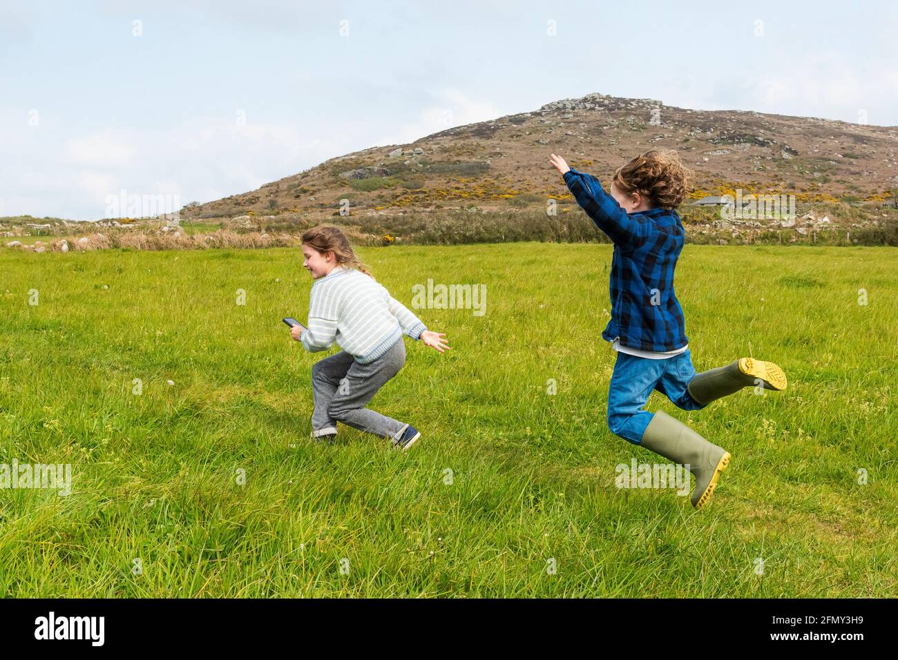 Excited energetic young children running through a field with Zennor Hill in the background in Cornwall. Stock Photo