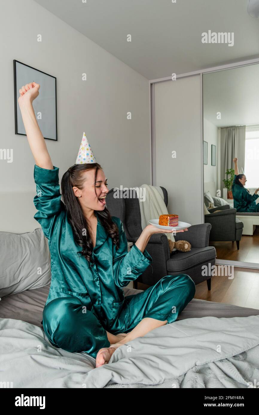 Young woman sits the bed her pajamas and holds a cake and screams with joy. Wellness concept. Rest at home Stock Photo