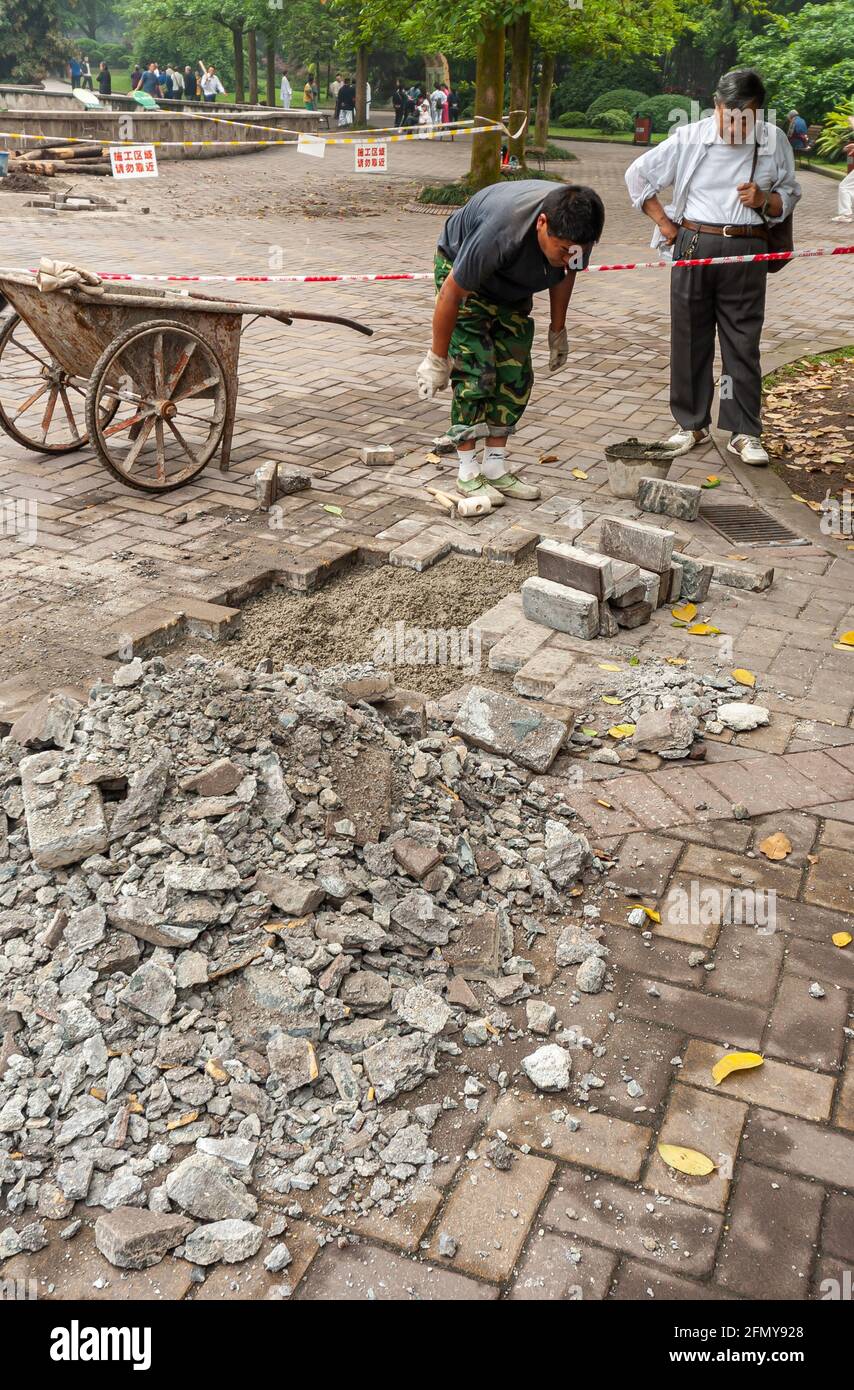 Chongqing, China - May 9, 2010: Downtown closeup. Male worker restores pavement with bricks while other man looks on. Wheelbarrow mearby. Park environ Stock Photo