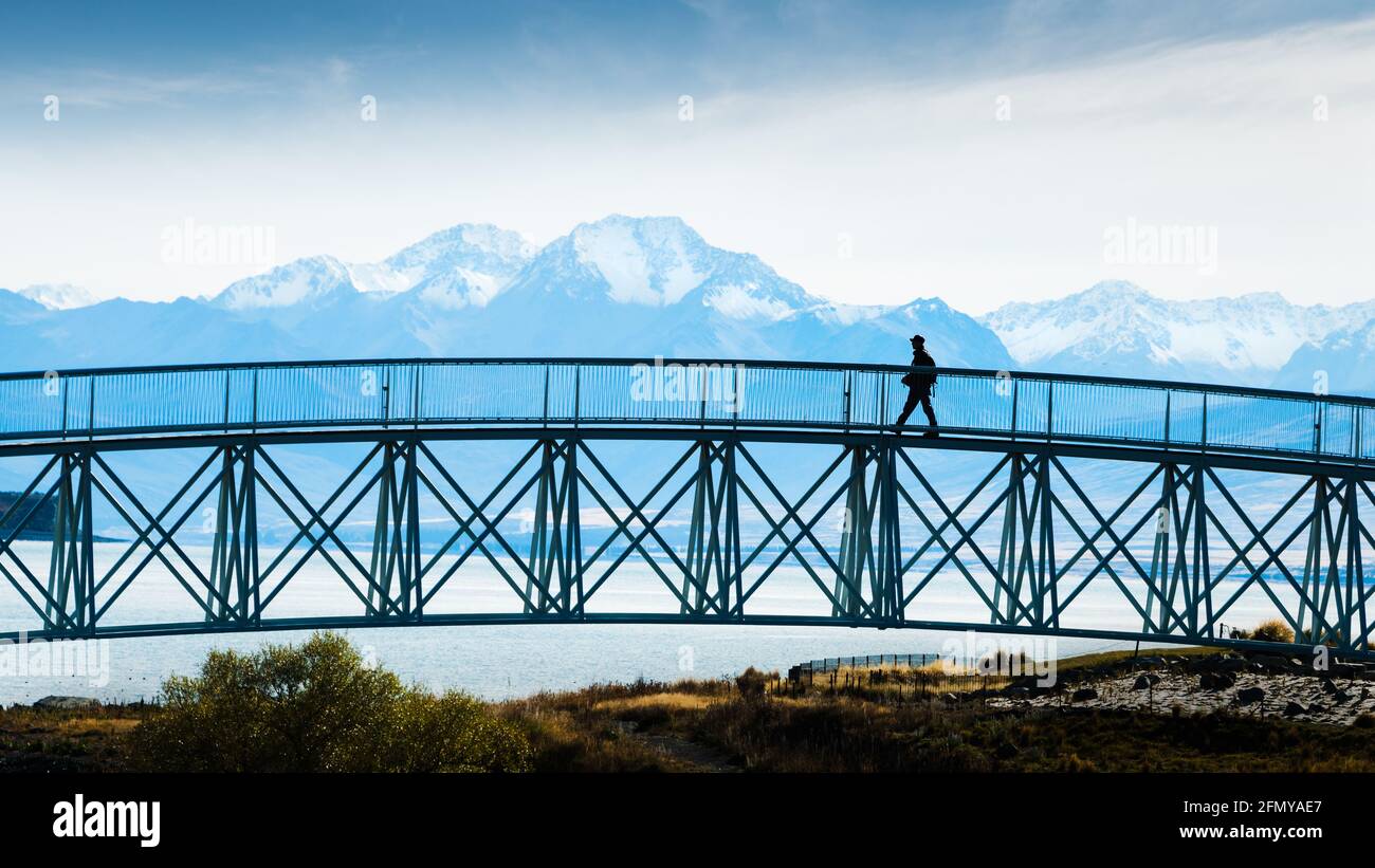 Tourist walking on the footbridge with mighty Southern Alps in the background, Lake Tekapo, South Island Stock Photo