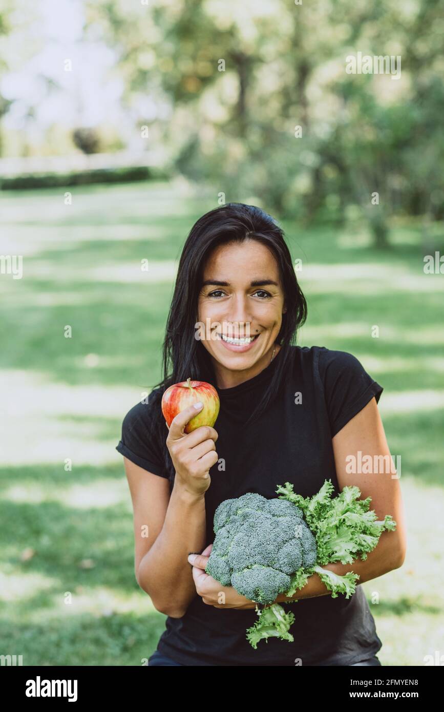 Portrait of smiling young tanned woman in the park holding an apple and a bouquet of greens with broccoli, diet, healthy eating. Soft selective focus. Stock Photo