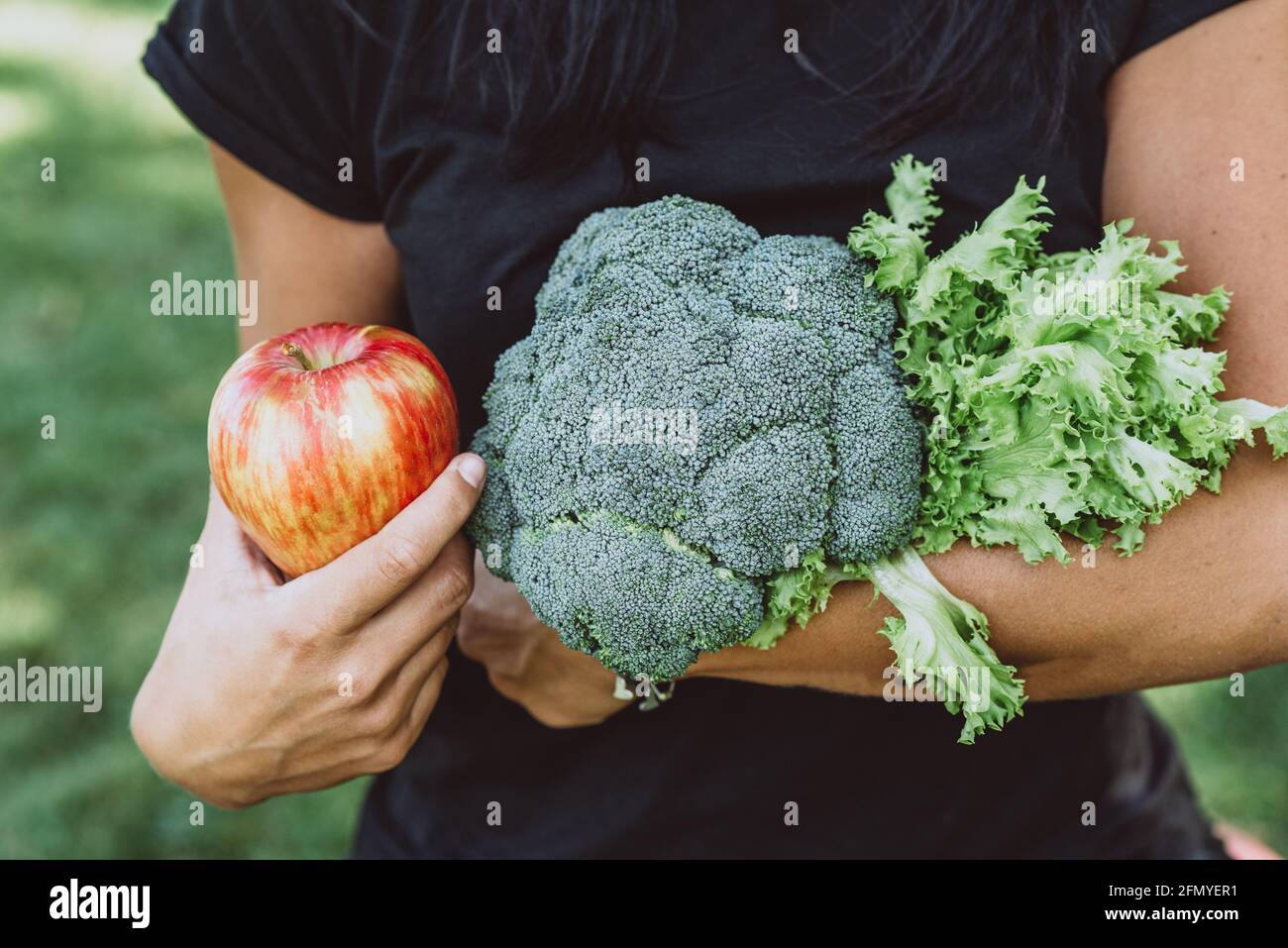 Portrait of tanned woman in the park holding an apple and a bouquet of greens with broccoli, diet, healthy eating. Soft selective focus. Stock Photo
