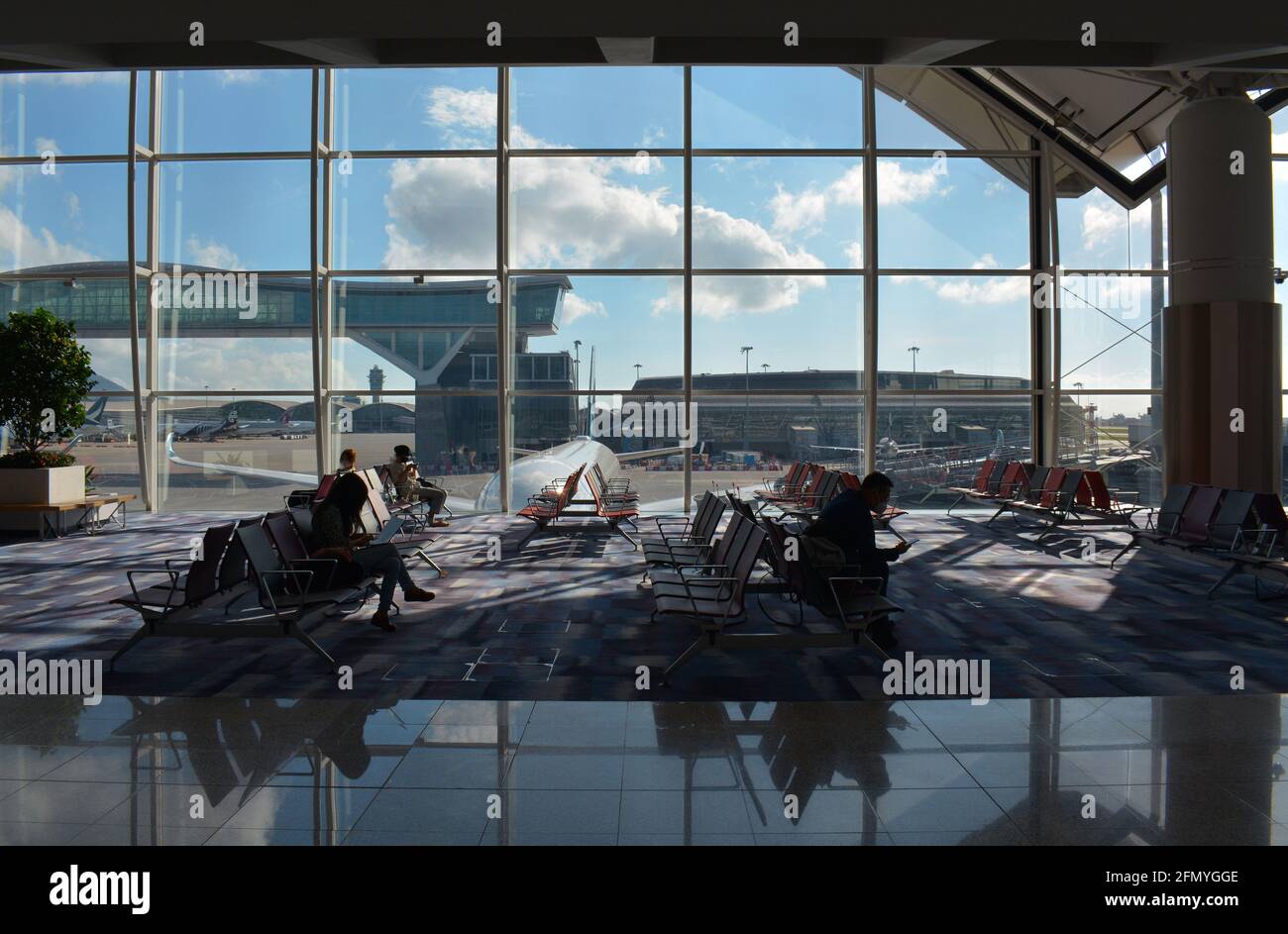 A few passengers wait for their flight in front of a huge window offering views of Chek Lap Kok, Hong Kong international airport. Stock Photo