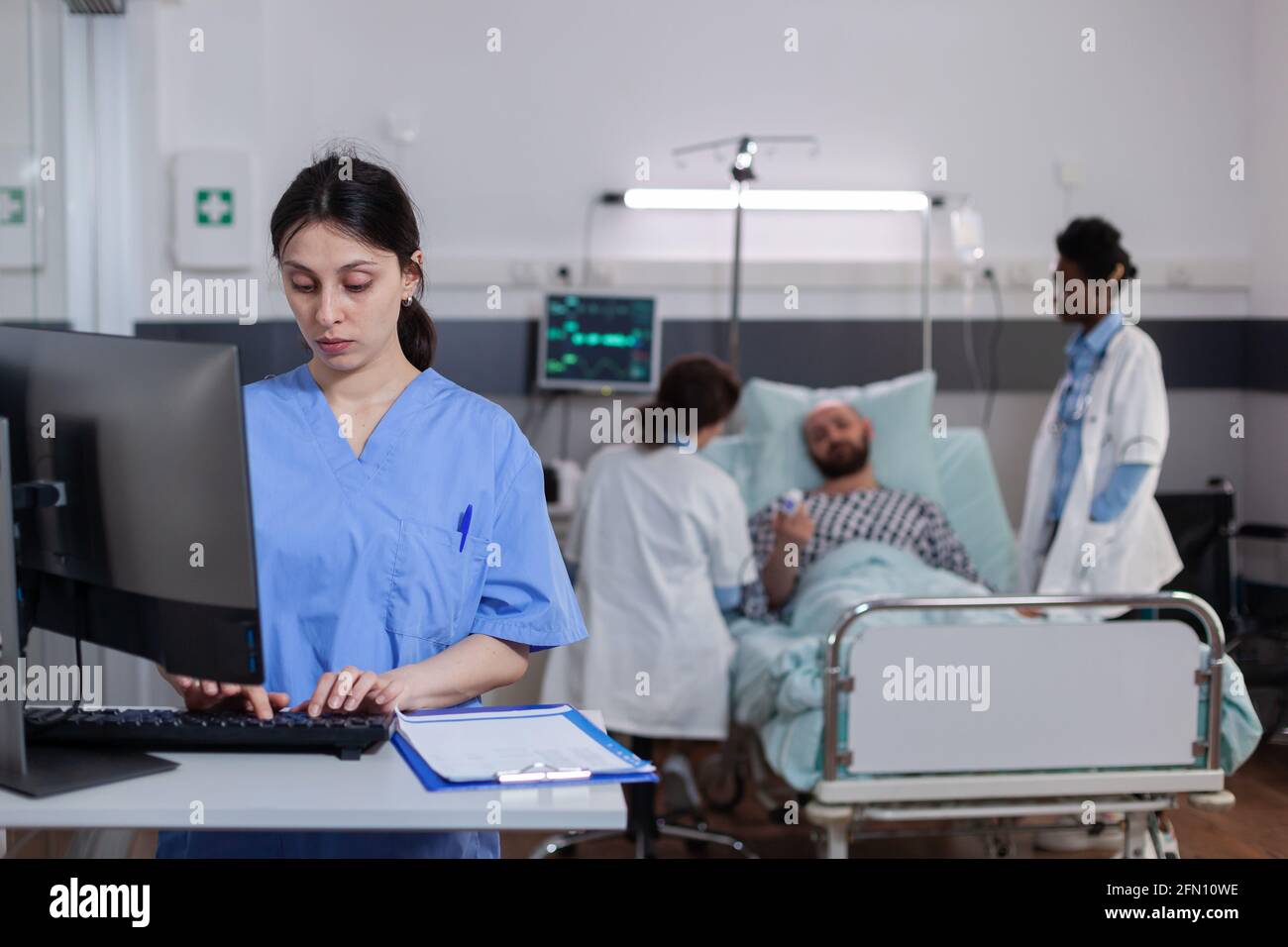 Woman asisstant typing medical recovery on computer while in background practitioner doctor monitoring sick man explaining disease symptom. Hospitalized patient having respiratory disorder Stock Photo