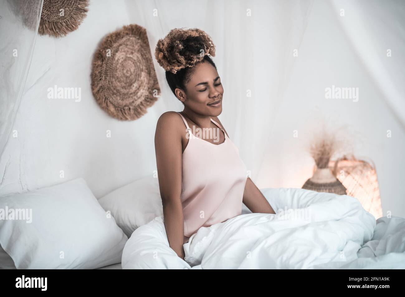 Joyful dark-skinned woman in nightie sitting on bed Stock Photo