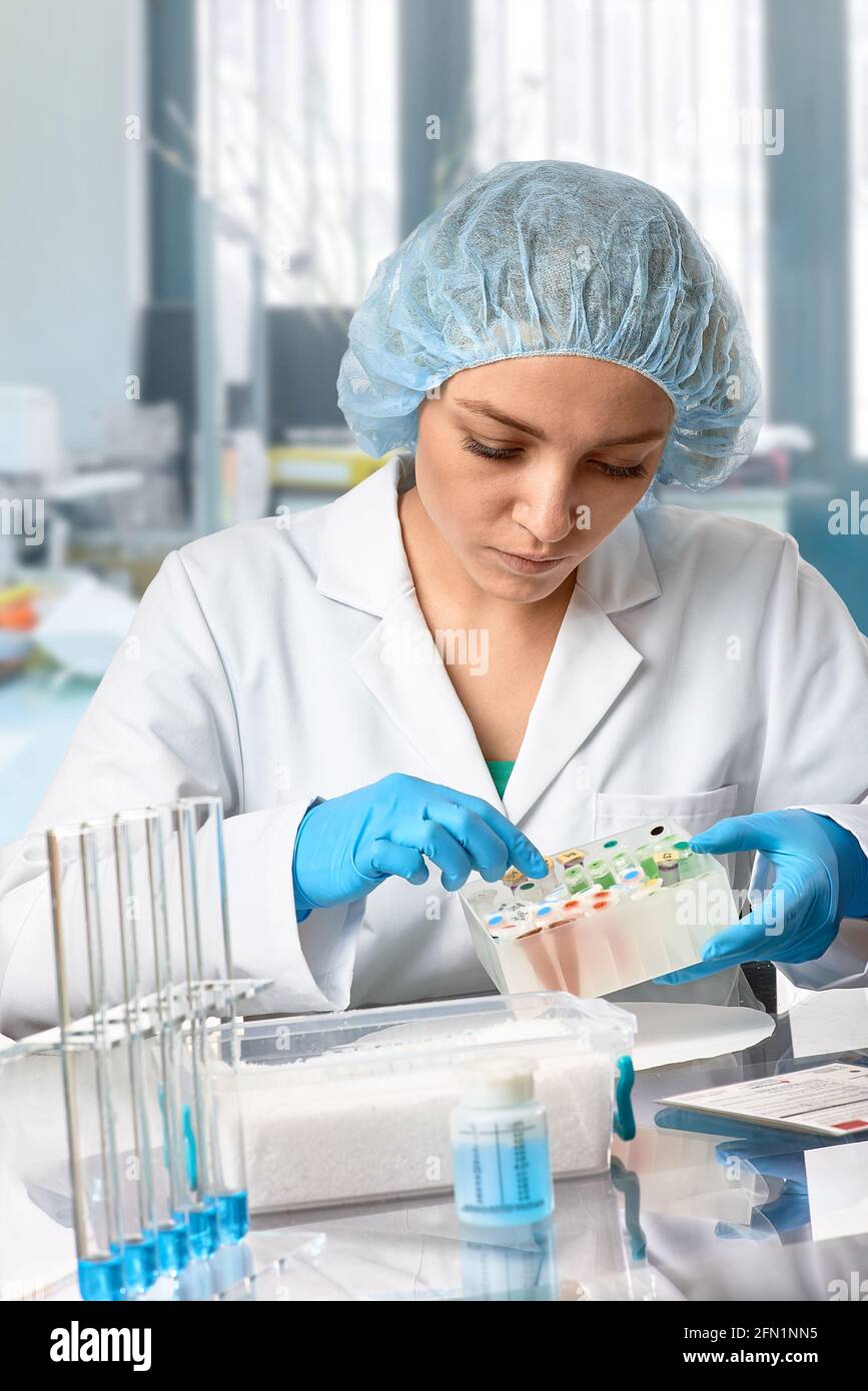 Pharma female tech works in laboratory. Caucasian young woman in protective gloves, hat and white gown pipettes sample in vial. Lab interior Stock Photo