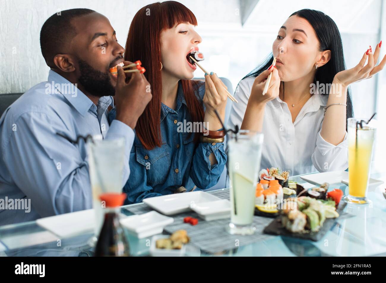 Lunch at a Asian restaurant. Close up shot of happy funny multiethnic  friends, black man and two pretty girls, eating sushi rolls with chinese  sticks Stock Photo - Alamy