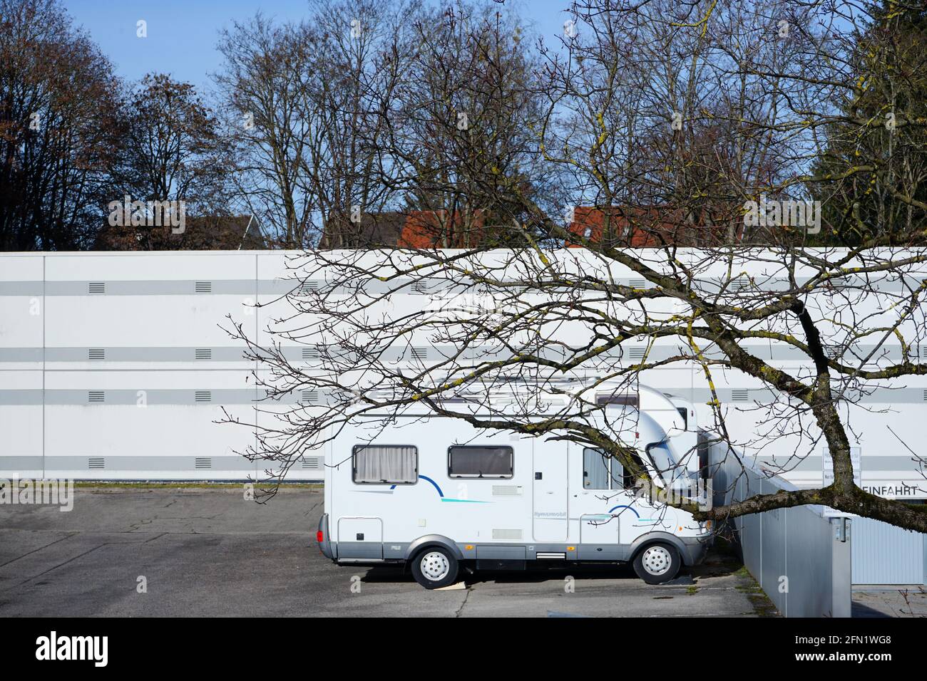 Parked camper in a parking lot in Munich, Westkreuz in front of a white noise barrier. In the foreground a tree with strong large branches. Stock Photo