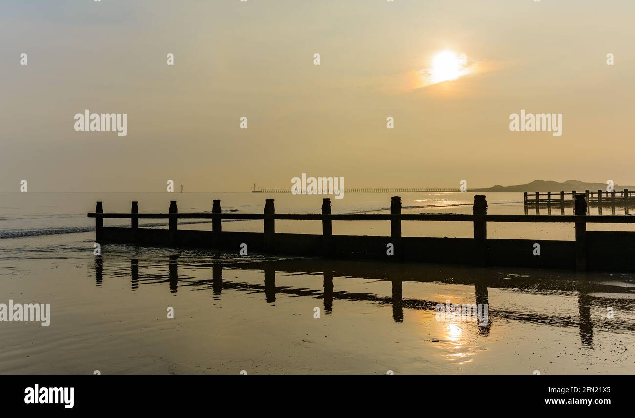 Calm sea at a sandy beach with a groyne and low sun, on a misty evening in West Sussex, England, UK. Stock Photo