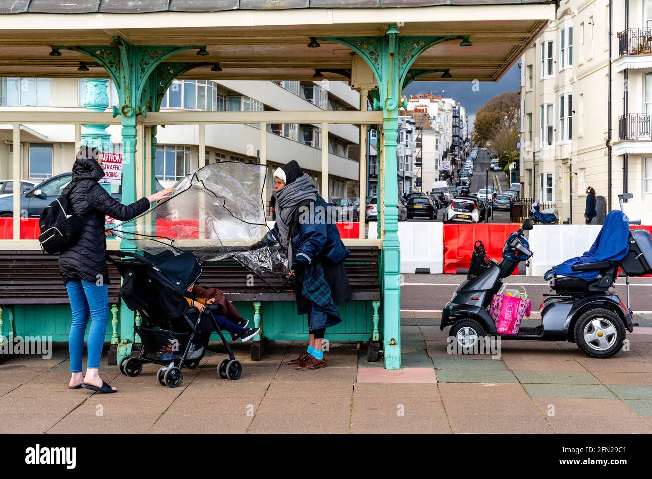 A Woman On A Mobility Scooter Stops To Help A Young Mother Put A Rain Cover Over A Baby In A Pushchair, Brighton, Sussex, UK. Stock Photo