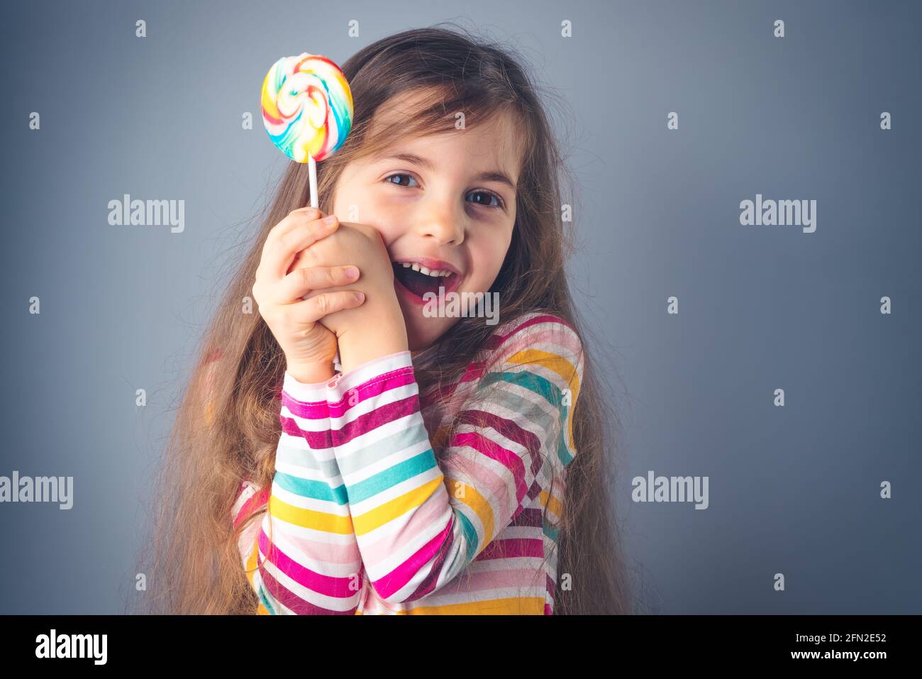 Little kid eating lollipop. Happy beautiful girl with candy isolated on blue background. Stock Photo