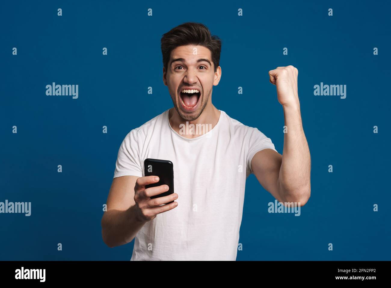 Delighted guy making winner gesture while using mobile phone isolated over blue background Stock Photo