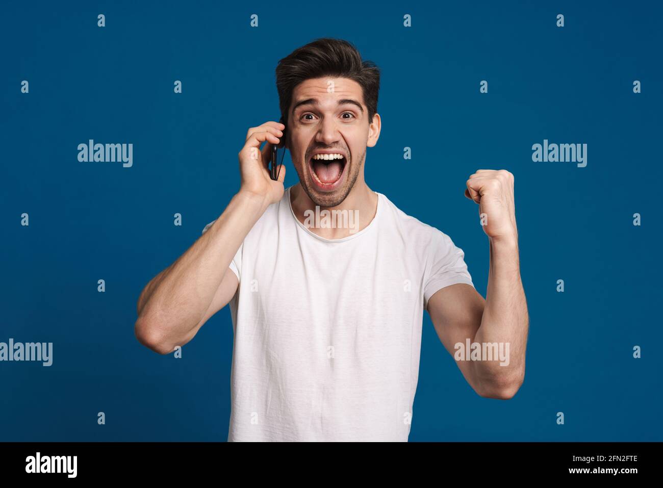 Delighted guy making winner gesture while talking on mobile phone isolated over blue background Stock Photo