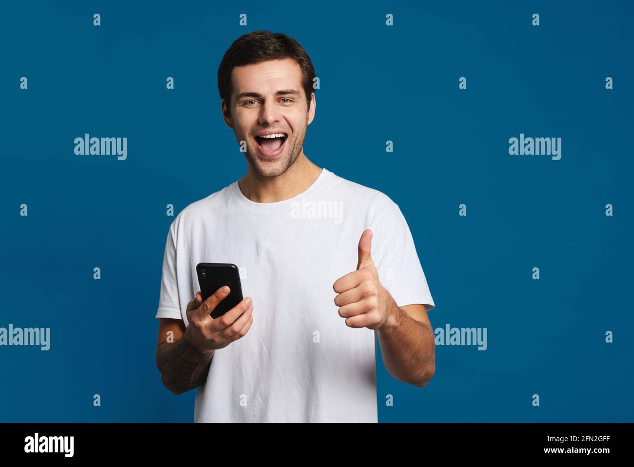 Happy unshaven guy showing thumb up while using mobile phone isolated over blue background Stock Photo