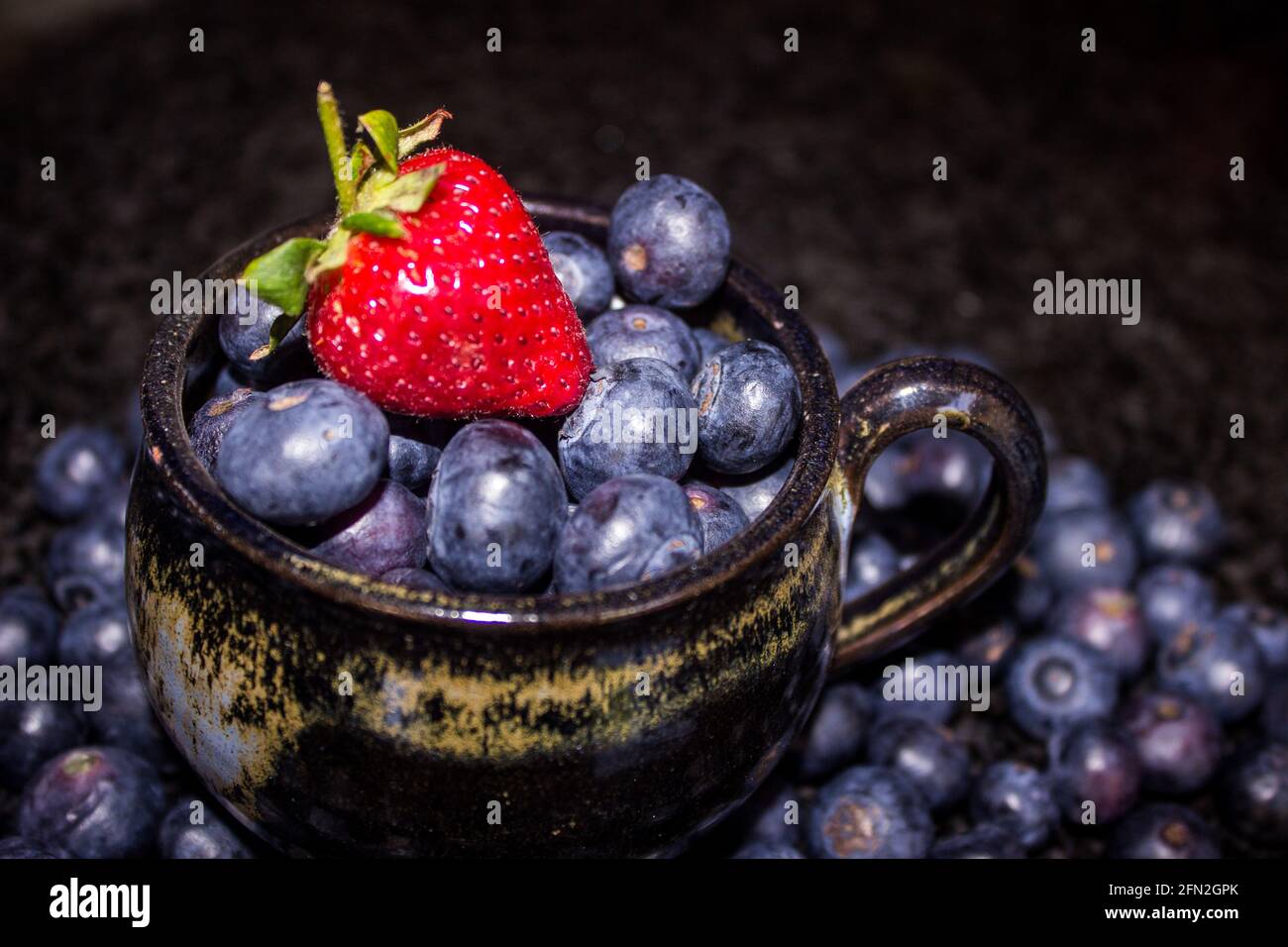 A single bright red Strawberry on top of a black tea-cup, filled with large indigo colored Blue berries Stock Photo