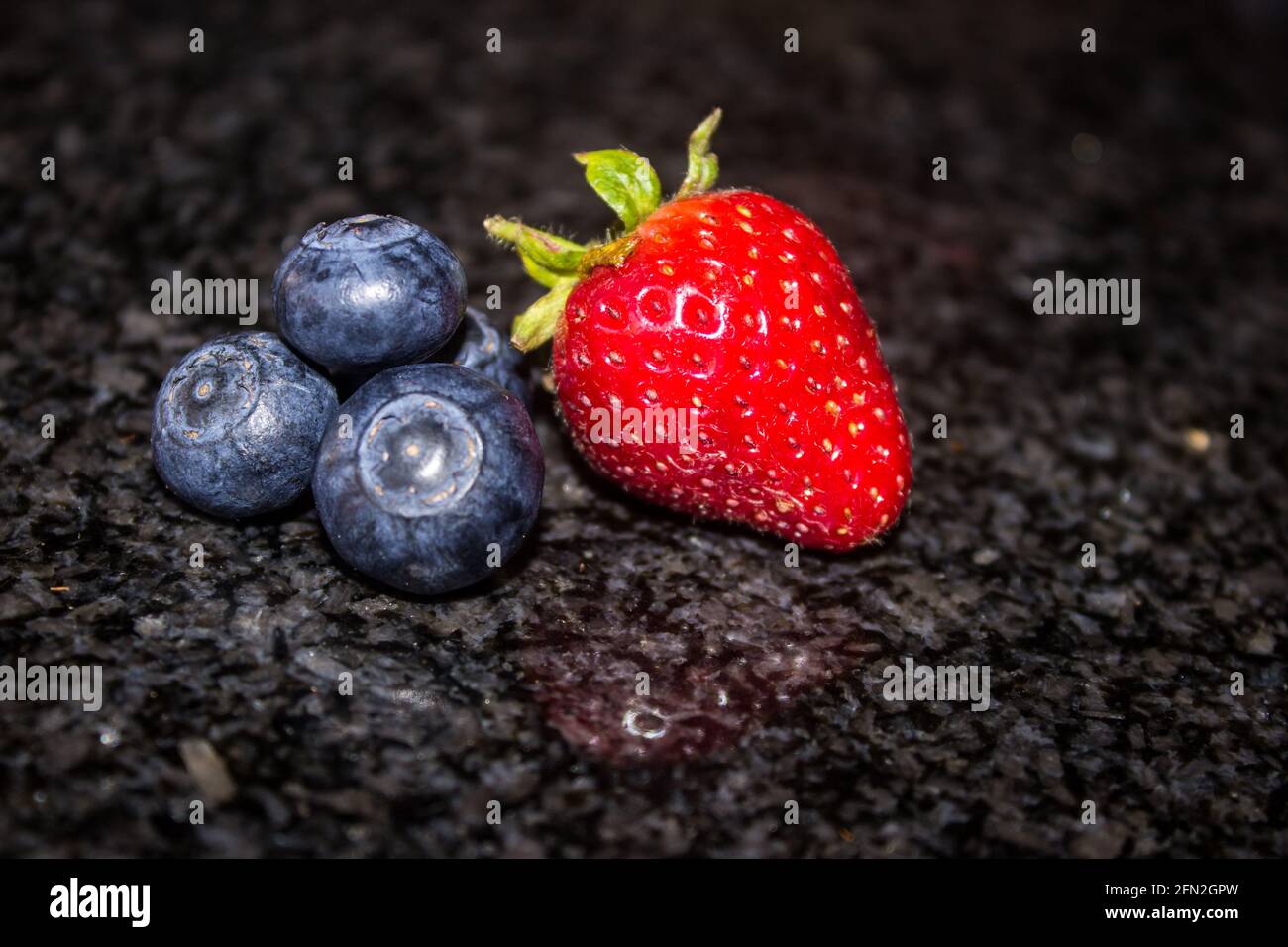 A single bright red strawberry and a small amount of indigo Blueberries on a dark kitchen countertop. Stock Photo