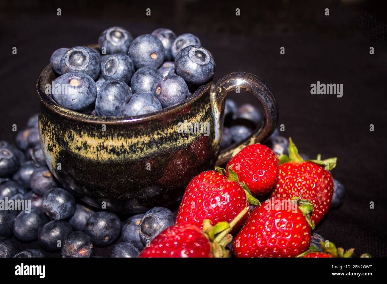 A black pottery teacup, filled to overflowing with blueberries, surrounded by strawberries and more blueberries Stock Photo