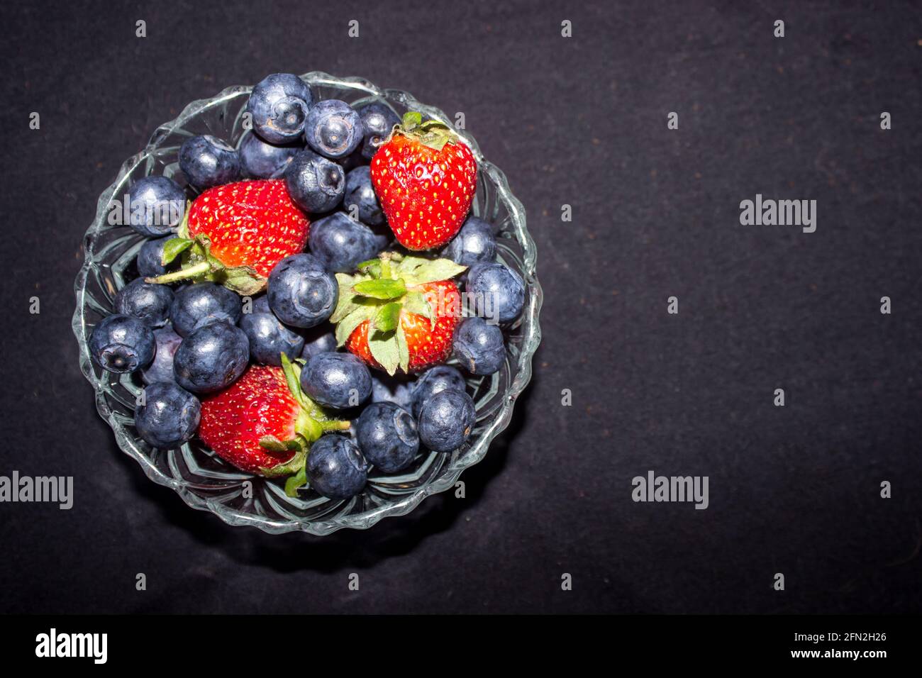 A small glass bowl filled with a mixture of blueberries and strawberries, against a black background Stock Photo